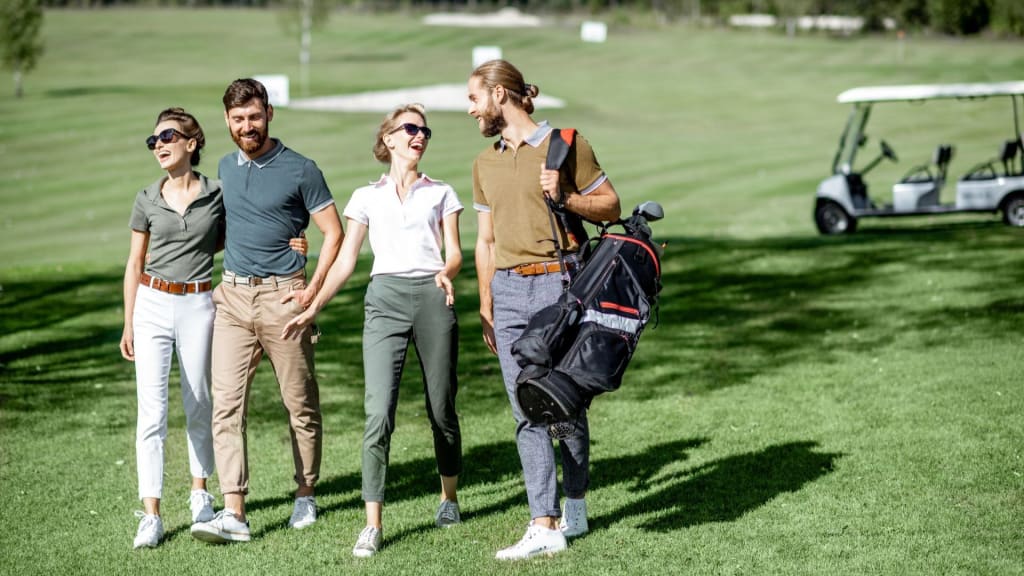 Four people walk together on a golf course, smiling and talking. One person carries a golf bag featuring top golf brands. A golf cart is visible in the background.